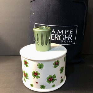May include: White ceramic diffuser base with a green four-leaf clover and ladybug design. The base is topped with a green metal burner. The text "Lampe Berger Paris" is printed on a black bag.