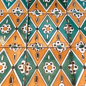 May include: Close-up of a collection of square ceramic tiles. Each tile features a repeating pattern of diamonds in teal and gold, with white floral and teardrop designs. The tiles are arranged in a grid, creating a cohesive decorative surface.