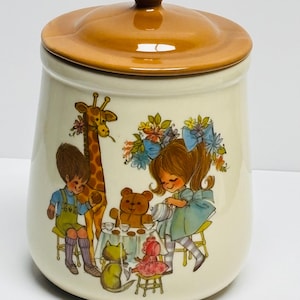 Puede incluir: Un tarro de cerámica con tapa marrón y cuerpo crema. El tarro presenta una ilustración caprichosa de niños y animales en una fiesta de té. La tapa y el pomo son de un tono marrón más oscuro.