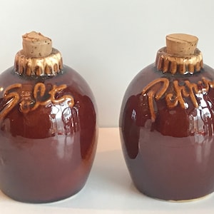 May include: A set of two brown ceramic salt and pepper shakers with cork stoppers. The shakers are shaped like round bottles and have the words "Salt" and "Pepper" written on them.