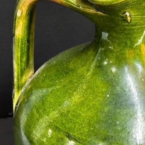 May include: A close-up of a green ceramic pitcher with a handle. The pitcher has a glossy finish with variations in the green color, from lighter to darker shades. The handle is arched and attached to the body of the pitcher.