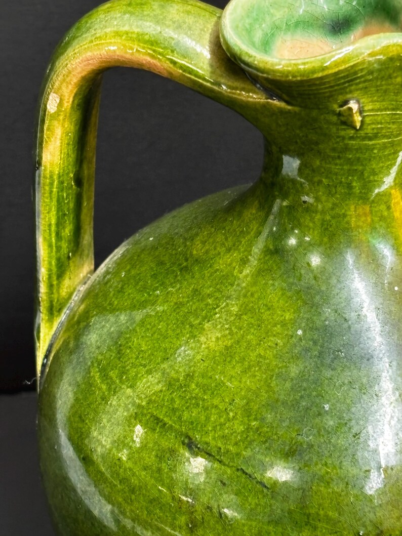 May include: A close-up of a green ceramic pitcher with a handle. The pitcher has a glossy finish with variations in the green color, from lighter to darker shades. The handle is arched and attached to the body of the pitcher.