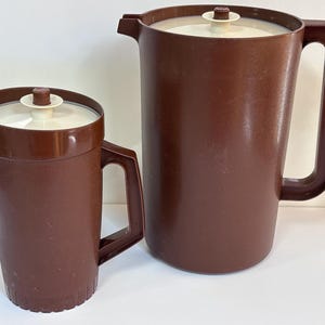 Può includere: Due caraffe in plastica marrone con coperchi color bianco sporco abbinati. Ogni caraffa presenta un manico laterale, un beccuccio e un pomello centrale sul coperchio. La caraffa più piccola ha una base scanalata, quella grande è liscia.