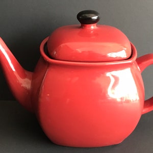 Op de afbeelding: Een rode theepot van keramiek met een zwarte knop op de deksel.