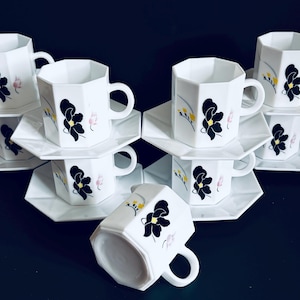 May include: A set of white ceramic cups and saucers. Each cup has a geometric design and is decorated with a black flower and yellow and pink accents. The saucers are white and have a geometric shape.