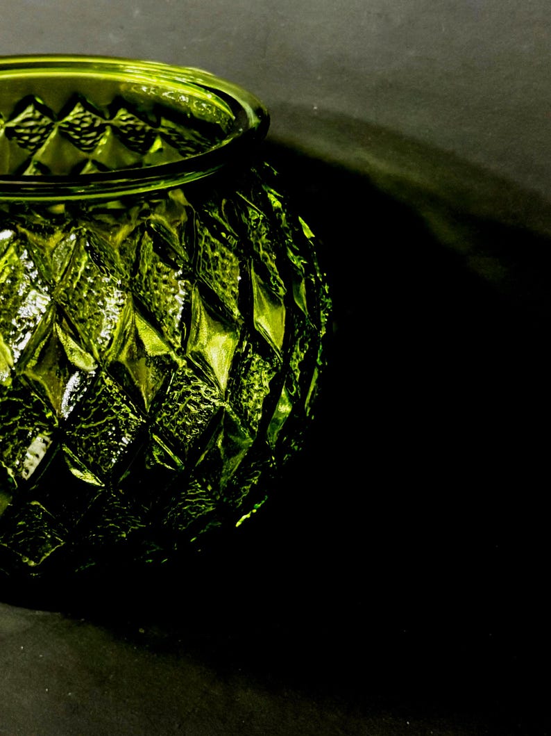 May include: A close-up of a green glass vase with a diamond pattern. The vase has a round shape with a wide opening and a textured surface. The glass reflects light, creating highlights and shadows.