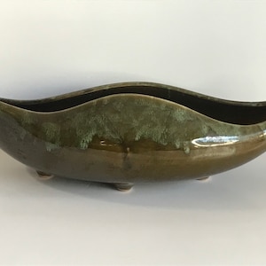 May include: A green ceramic planter with a unique, wave-like shape and four small feet. The planter has a mottled green glaze with a darker green rim.