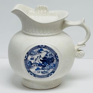 May include: A white ceramic jug with a lid and handle. The jug features a blue and white circular design with a landscape scene. The jug has a speckled texture and a spout for pouring.