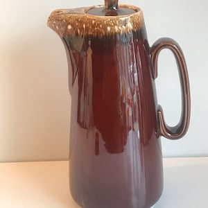 以下が含まれることがあります： 蓋付きの茶色の釉薬をかけたセラミック製のコーヒーポット。ポットは茶色と白の釉薬で、まだら模様になっています。