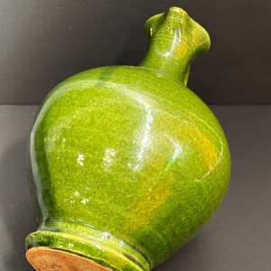 May include: A green ceramic vase with a bulbous body and a flared, ruffled neck. The vase has a glossy finish with variations in the green color. The base of the vase is a light brown color.