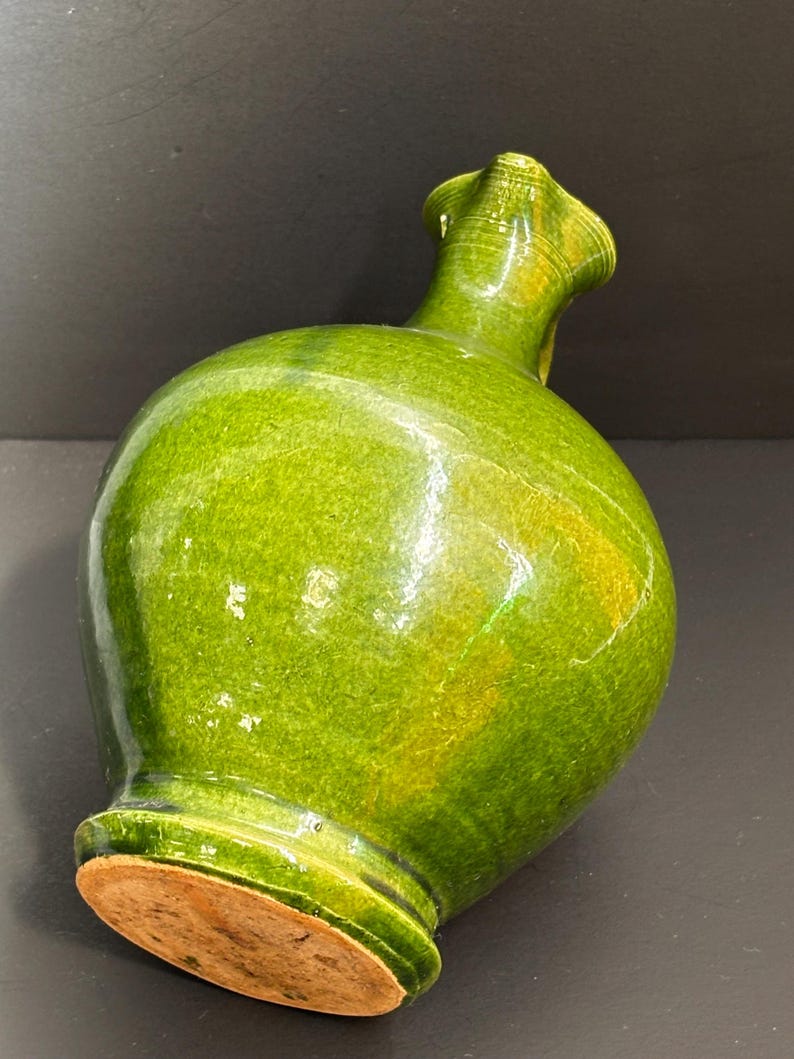 May include: A green ceramic vase with a bulbous body and a flared, ruffled neck. The vase has a glossy finish with variations in the green color. The base of the vase is a light brown color.