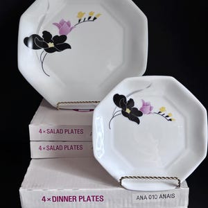 May include: Two white octagonal plates featuring a floral design with black, purple, and yellow petals. The plates are displayed on white boxes labeled 4x SALAD PLATES and 4x DINNER PLATES with the text ANA 010 ANAIS printed on the side labels.