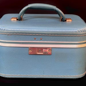 May include: A light blue rectangular vanity case with a textured exterior, a top handle, and a metallic latch closure. The case features white piping trim along the edges and a central silver-toned locking mechanism on the front panel.