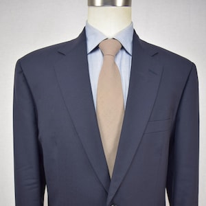 May include: A navy blue suit jacket with a light blue dress shirt and a tan tie. The jacket has two front pockets and a single-button closure. The suit is displayed on a mannequin.
