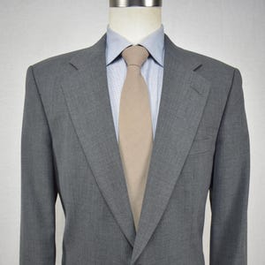 May include: A gray suit jacket with a light blue dress shirt and a tan tie. The jacket has two buttons and two front pockets. The suit is displayed on a mannequin.