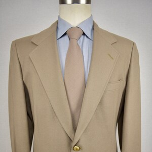 May include: A tan blazer with a single gold button. The blazer has a notched lapel and a pocket on the left side. The blazer is worn over a light blue shirt with a tan tie.