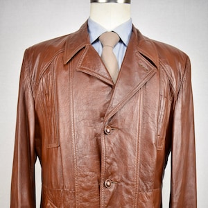 Puede incluir: Una chaqueta de cuero marrón con cierre de botones y cuello. La chaqueta tiene una superficie ligeramente texturizada y está diseñada con un estilo clásico.