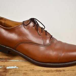 May include: A single brown leather dress shoe with dark brown laces. The shoe has a classic Oxford style with a rounded toe and a low heel. The leather appears smooth and polished, with visible stitching details. The shoe is set against a neutral background.