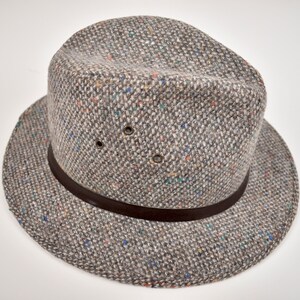 May include: A grey fedora hat crafted from a textured tweed fabric featuring small flecks of blue, orange, and white throughout. The hat includes a dark brown leather band encircling the base of the crown and two metal ventilation eyelets on the side.