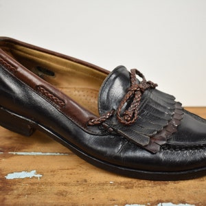 May include: Black leather loafer with brown leather detailing. The shoe has a braided leather feature and fringe on the vamp. The sole is black, and the shoe is on a wooden surface.