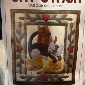 May include: A wall quilt kit featuring a stack of three cats with hearts and leaves. The kit includes all fabrics, woolfelt, patterns, instructions, batting, buttons, and embroidery floss. The quilt measures 13 inches by 15 inches.