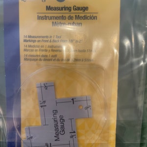 Puede incluir: Una regla de medición de plástico blanco con marcas en pulgadas y fracciones de pulgada. La regla tiene forma de T y tiene marcas en ambos lados. La regla está empaquetada en una bolsa de plástico transparente con una etiqueta amarilla y blanca que dice "Dritz Quilting Measuring Gauge Instrumento de Medición Mètre-ruban". La etiqueta también incluye instrucciones en inglés, español y francés.