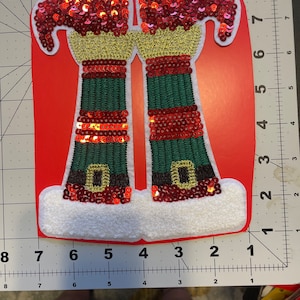 Puede incluir: Un par de piernas de elfo verdes y rojas con ribetes dorados y lentejuelas rojas. Las piernas están unidas a una base blanca y peluda.