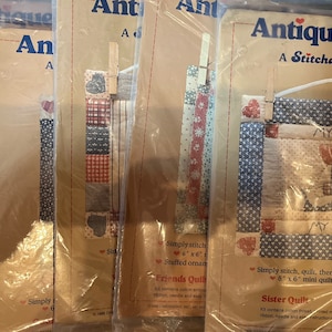 Może przedstawiać: Pięć pakietów z zestawami tkanin do tworzenia pikowanych wiszących ozdób ściennych. Pakiety są oznaczone "Antiqued Quilts, A Stitchables Collection". Jeden pakiet zawiera projekt z dwoma postaciami z tekstem "My Sister, My Friend".