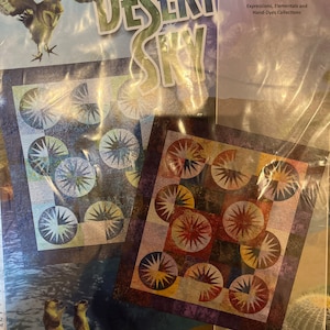 May include: A quilting pattern featuring a desert sky theme with a blue and brown color scheme. The pattern includes a variety of starburst shapes and circles. The pattern is titled "Desert Sky" and is by Riley Blake Designs.