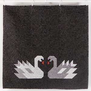May include: A black and gray quilt featuring two stylized swans facing each other. The swans are composed of geometric shapes in shades of gray and white, with red beaks. The background is a textured black fabric.