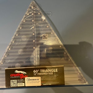 May include: A clear plastic 60 degree triangle ruler with black markings. The ruler is 12 inches in size and is labeled "Creative Grids 60° Triangle 12" Finished Size".