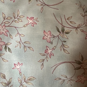 May include: Pale blue fabric with a floral design. The pattern includes pink flowers, green leaves, and brown stems. The fabric has the text "by Maywood Studio" and "www.MaywoodStudio.com" printed on it.