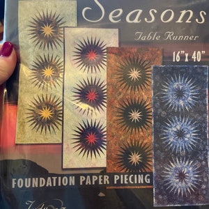 Pode incluir: Um padrão de acolchoado para um corredor de mesa chamado "One For All Seasons" de Judy Niemeyer Quilting. O padrão apresenta três designs diferentes de explosões de estrelas em tons de azul, vermelho e amarelo. O padrão é para o acolchoado de papel de fundação e mede 40,6 cm por 101,6 cm.
