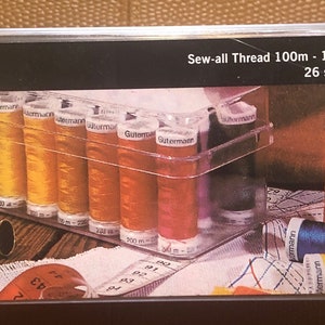 Puede incluir: Una caja de plástico transparente llena de 26 carretes de hilo Gütermann Sew-all, 100 m cada uno, 100 % poliéster. La caja está etiquetada con "Sew-all Thread 100m - 100% polyester 26 spools / shades" y "Gütermann Sewing Thread Box".