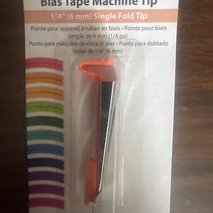 May include: A Simplicity brand bias tape machine tip in a 1/4 inch single fold size. The tool features a metallic silver metal guide attached to an orange plastic component, encased in clear plastic packaging with a white card backing.