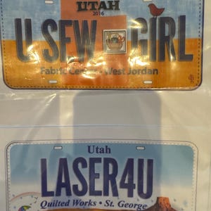 Könnte beinhalten: Zwei dekorative Nummernschilder. Das obere Schild lautet "U SEW GIRL" mit "Utah 2016" und "Fabric Center West Jordan". Das untere Schild lautet "LASER4U" mit "Quilted Works St. George" und einer Wüstenlandschaft.