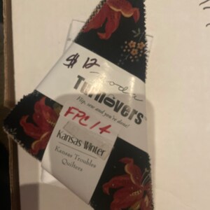 May include: A black fabric with a red floral pattern. The fabric is labeled "Turnovers" and "FPC 14" with the text "Kansas Winter" and "Kansas Troubles Quilters".