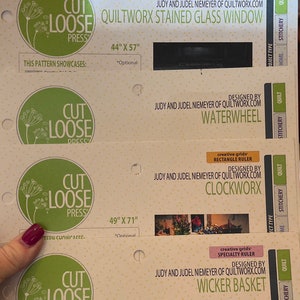 May include: A close-up of a product display for quilting patterns. The display features five different patterns, each with a sample quilt block, the pattern name, and the designer's name. The patterns are: Labyrinth, Quiltworx Stained Glass Window, Waterwheel, Clockworx, and Wicker Basket.