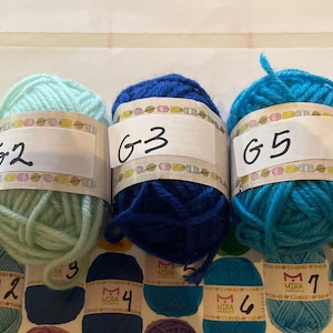May include: Five balls of yarn in different colors: green, light blue, dark blue, turquoise, and light blue. Each ball is labeled with a letter G and a number. The labels are white with black lettering.