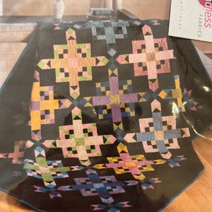 May include: A black quilt with a geometric pattern in shades of pink, yellow, green, blue, and purple. The quilt features a repeating cross-like design with square and triangular elements. The quilt is displayed on a wooden surface.