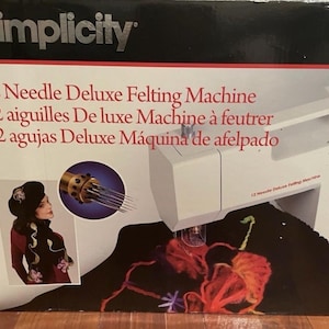 May include: A white Simplicity 12 Needle Deluxe Felting Machine box featuring a graphic of the machine, a close-up of the needle mechanism, and a person wearing a black beret and scarf. The text reads 12 Needle Deluxe Felting Machine.
