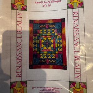 May include: A paper pieced stained glass wall hanging titled "Renaissance Beauty." The design features a central floral motif surrounded by geometric shapes in shades of red, blue, yellow, and green. The dimensions are 34 x 46 inches.