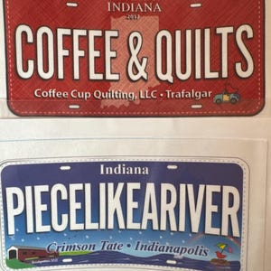 May include: Two decorative license plates. The top plate is red with white text that reads "COFFEE & QUILTS" and "INDIANA 2017". The bottom plate is blue with white text that reads "PIECE LIKE A RIVER" and "INDIANA".