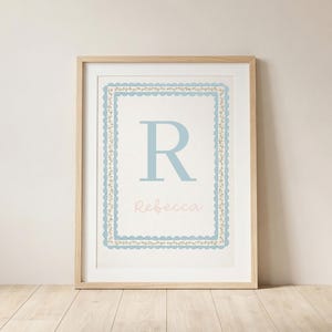 May include: A framed print featuring a light blue letter "R" and the name "Rebecca" in a soft pink font. The artwork is surrounded by a decorative blue and gold border. The frame is made of light-colored wood, and the background is a neutral color.