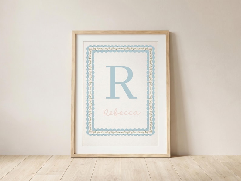 May include: A framed print featuring a light blue letter "R" and the name "Rebecca" in a soft pink font. The artwork is surrounded by a decorative blue and gold border. The frame is made of light-colored wood, and the background is a neutral color.