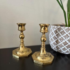 Puede incluir: Dos candelabros antiguos de latón con bases hexagonales y detalles ornamentados. Los candelabros tienen un tono dorado cálido y están colocados sobre una superficie oscura. Una maceta decorativa con estampado blanco y marrón con una planta verde está en el fondo.