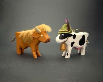 Puede incluir: Dos figuras de animales de madera. Una es una vaca marrón con una melena beige esponjosa y cuernos pequeños. La otra es una vaca blanca con manchas negras, que lleva un sombrero de fieltro verde con una pluma y una campana dorada al cuello.