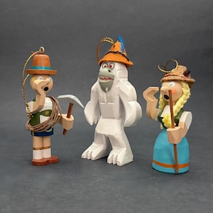 Puede incluir: Tres adornos de madera que incluyen una figura con chaleco verde, pico y cuerda, un yeti blanco con sombrero naranja y pluma azul, y una figura con falda azul, trenzas rubias y sombrero marr&oacute;n, cada uno con un lazo dorado para colgar.