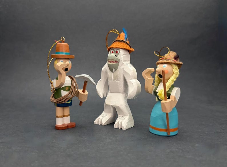 Puede incluir: Tres adornos de madera que incluyen una figura con chaleco verde, pico y cuerda, un yeti blanco con sombrero naranja y pluma azul, y una figura con falda azul, trenzas rubias y sombrero marr&oacute;n, cada uno con un lazo dorado para colgar.