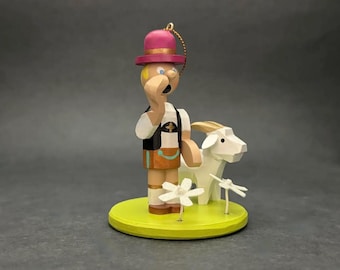 May include: A wooden figurine of a person wearing a pink hat, white shirt, black vest, and brown shorts, standing on a yellow circular base. Beside the figure is a white goat and two white flowers. A gold hanging cord is attached to the hat.